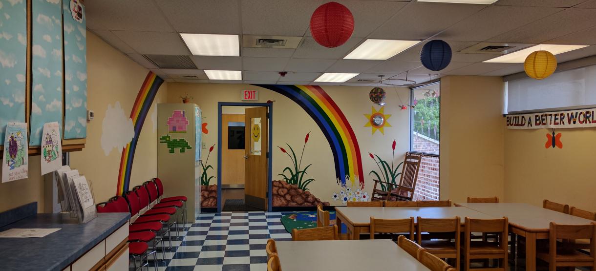 Picture of children's craft room with rainbow painted on the wall, several tables and chairs.